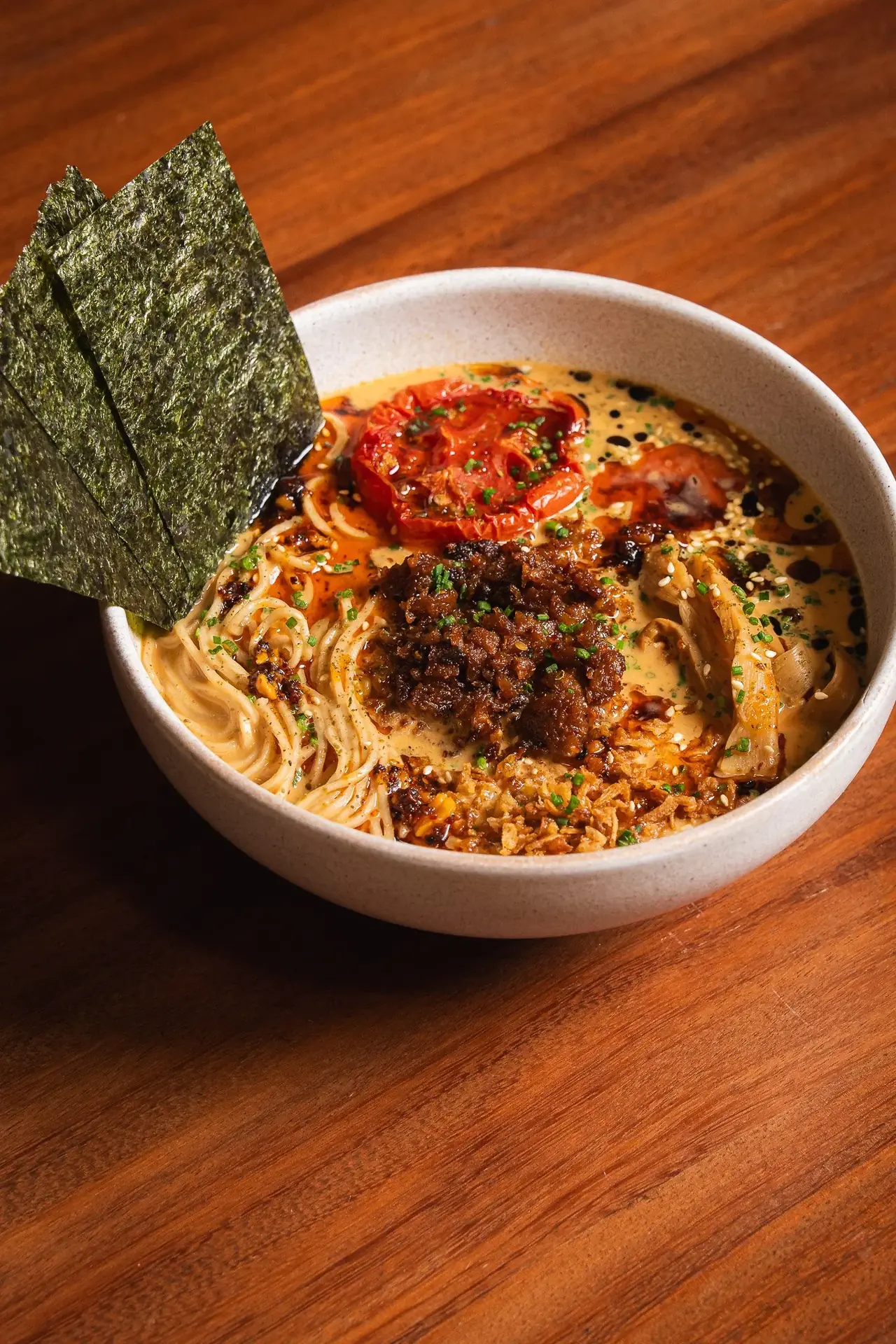 Bowl of noodle ramen in a creamy broth topped with crispy nori sheets, a roasted tomato half, chili crisp, and mushrooms, at Umami Plant Based