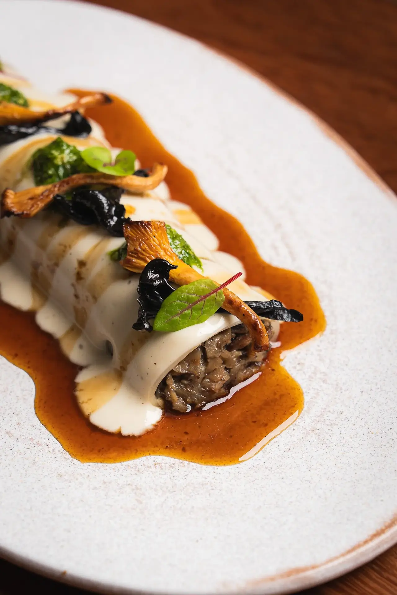 Rolled dish with a creamy sauce, orange-tinted jus, sauteed mushrooms, black fungus, and herb garnish on an oval plate, at Umami Plant Based