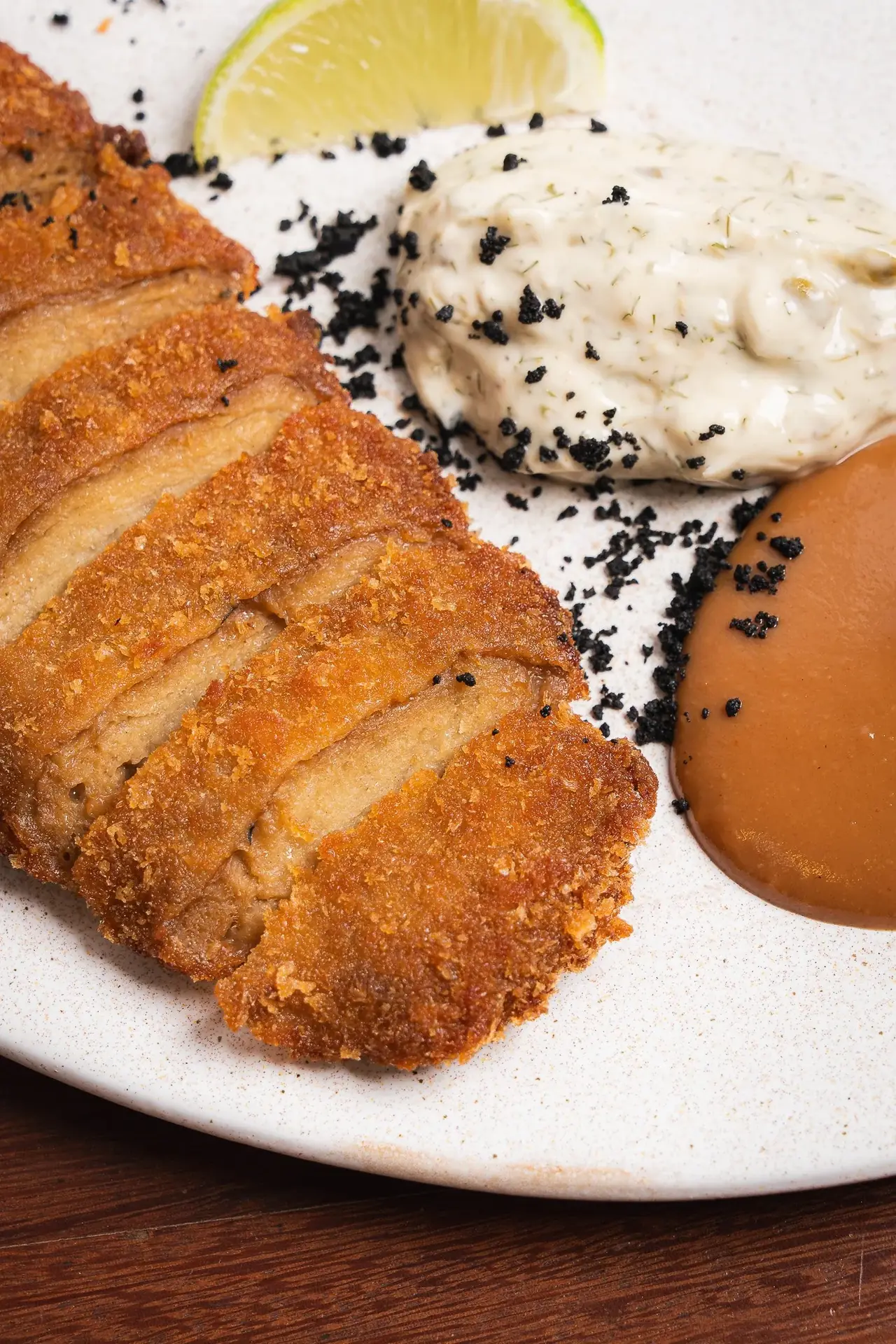 Close-up of sliced breaded and fried cutlet with a lime wedge, black sesame-flecked tartar sauce, and a brown dipping sauce, at Umami Plant Based