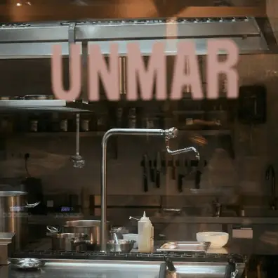 The restaurant's open kitchen seen through the storefront window, with pots on the stove, a knife rack and squeeze bottles, and the UNMAR name lettered on the glass