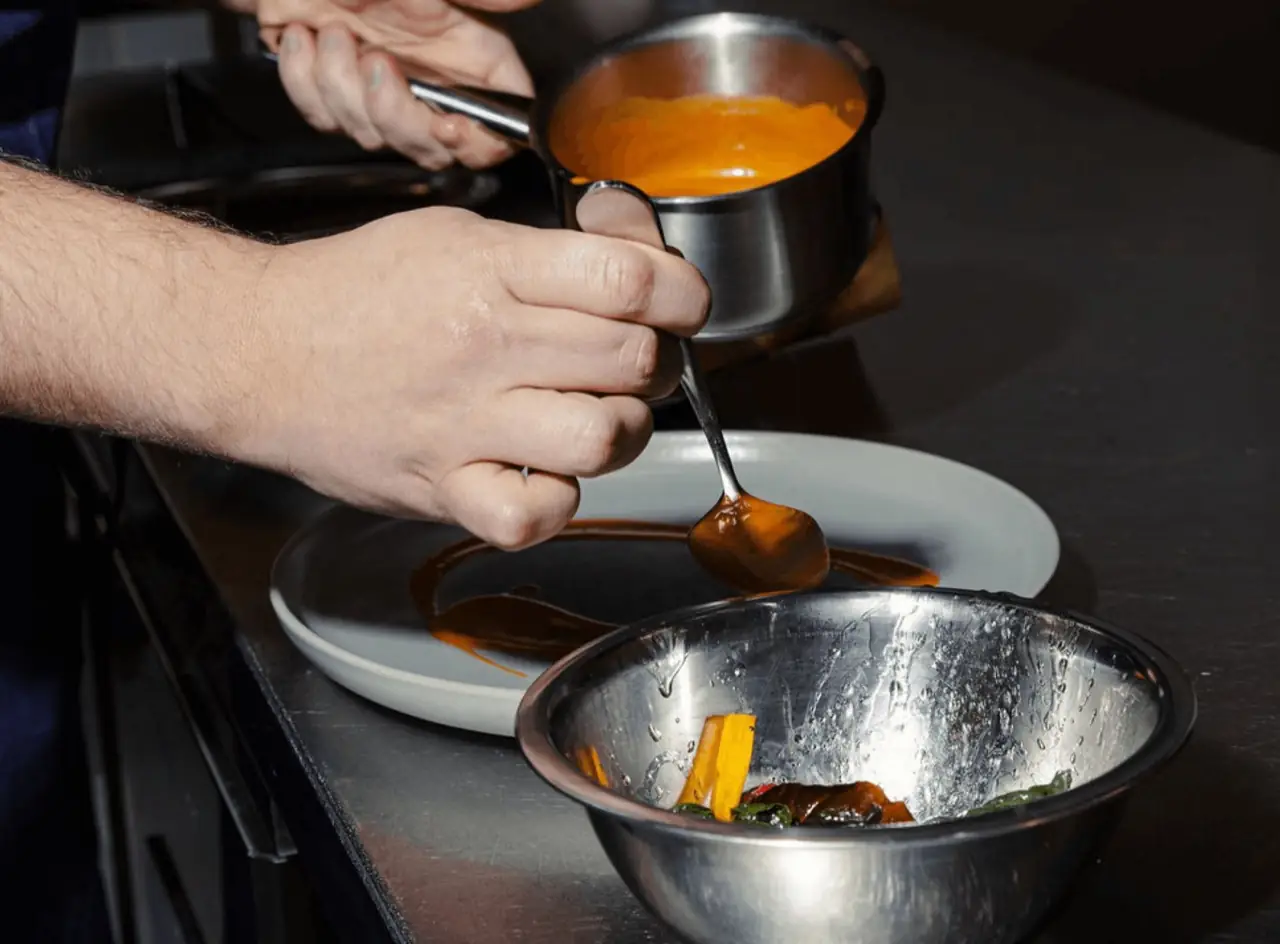 A cook's hands spooning an orange sauce from a small saucepan onto a white plate beside a metal mixing bowl of roasted peppers