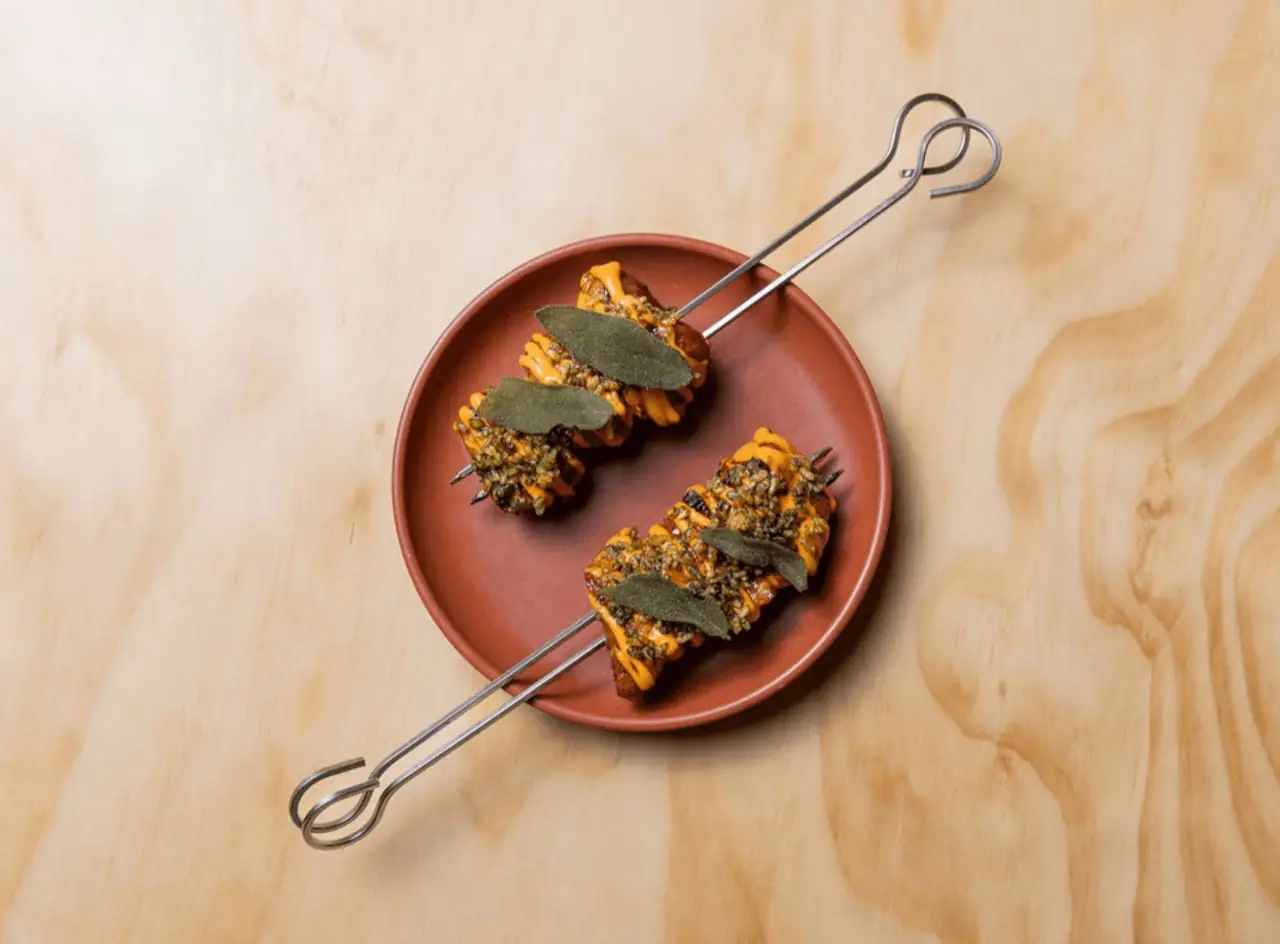 Two metal skewers of sauced, seed-crusted morsels topped with fried sage leaves on a terracotta plate