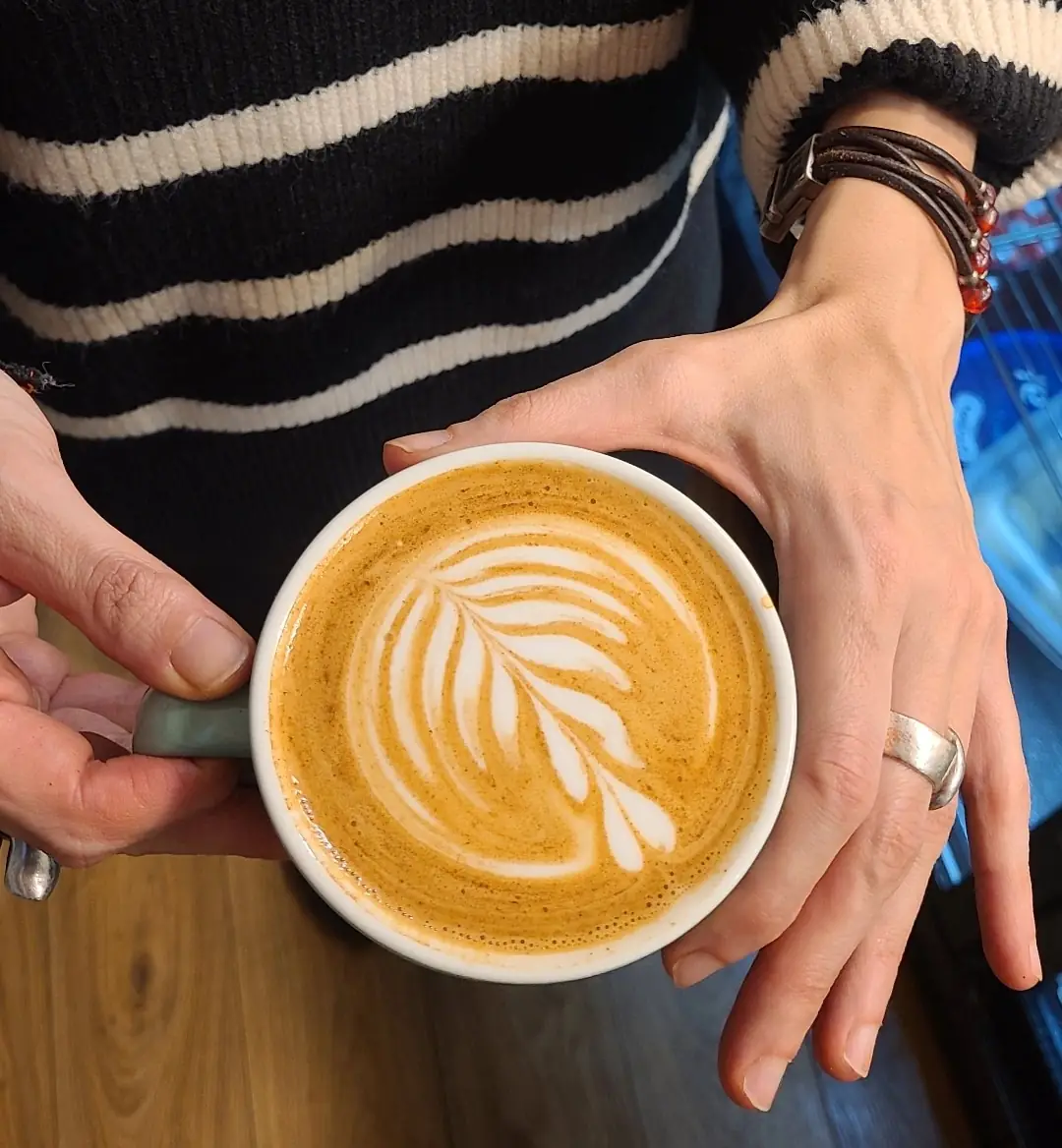 A latte with fern latte art in a ceramic cup held in two hands over a wooden table
