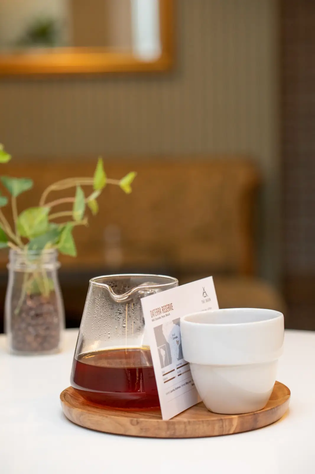 A glass carafe of filter coffee, a white cup and a tasting card on a round wooden board on a café table