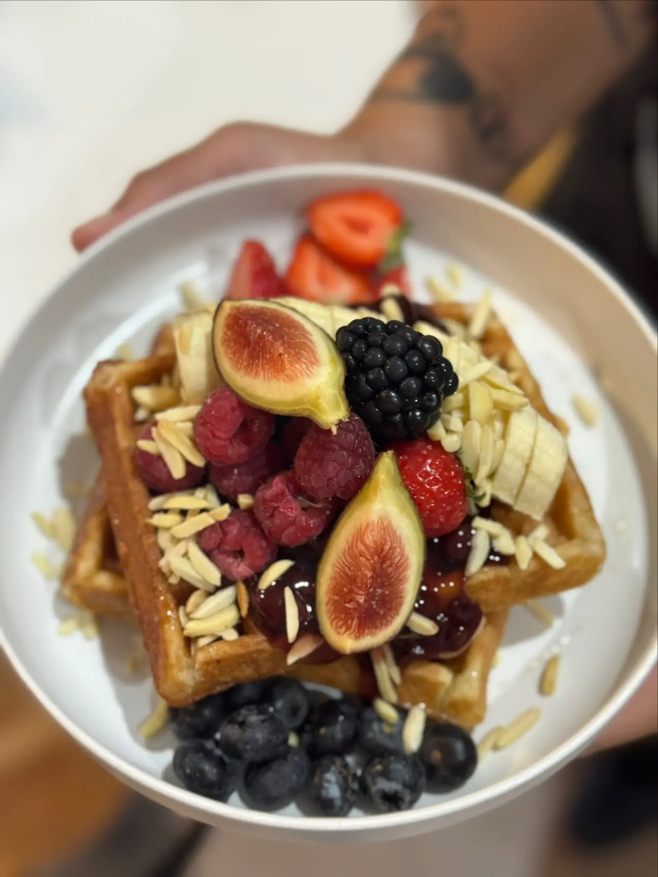 A waffle topped with figs, blackberries, raspberries, banana slices, blueberries, and sliced almonds.