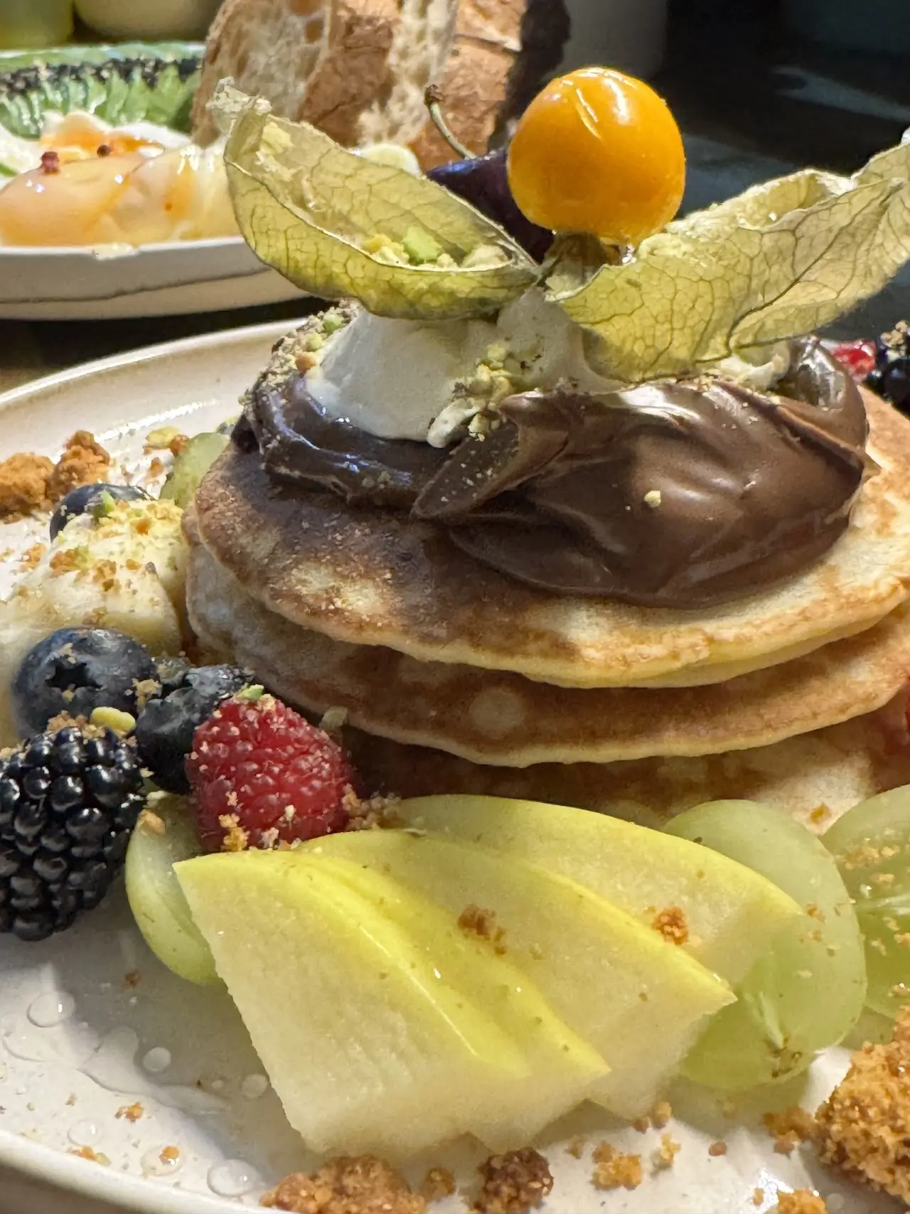 A stack of pancakes topped with chocolate spread, served with a side of berries and sliced pear.