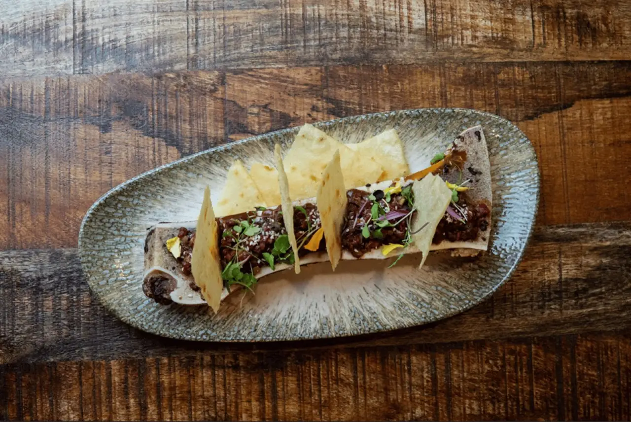 Roasted bone marrow topped with beef tartare, sesame, microgreens and crisp cracker shards on an oval speckled plate over reclaimed wood