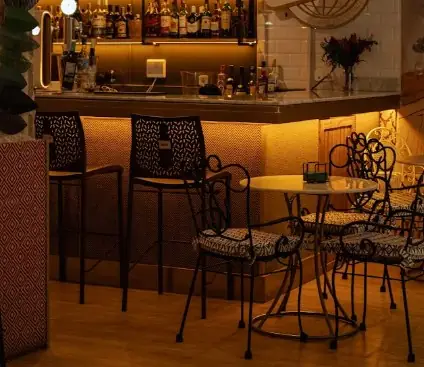 Warmly lit bar interior with backlit bottle shelves, patterned bar stools and wrought-iron cafe tables and chairs on a wood floor