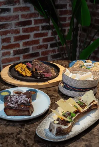 Spread of dishes on a marble table: sliced grilled steak on a round board, glazed ribs with sauces, a basket of bread and bone marrow with cheese