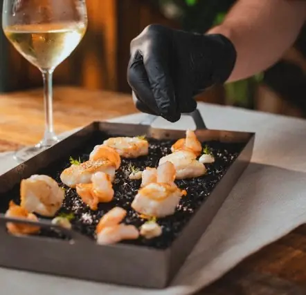 Grilled prawns on a black slate tray beside a glass of white wine, a gloved hand plating with tongs