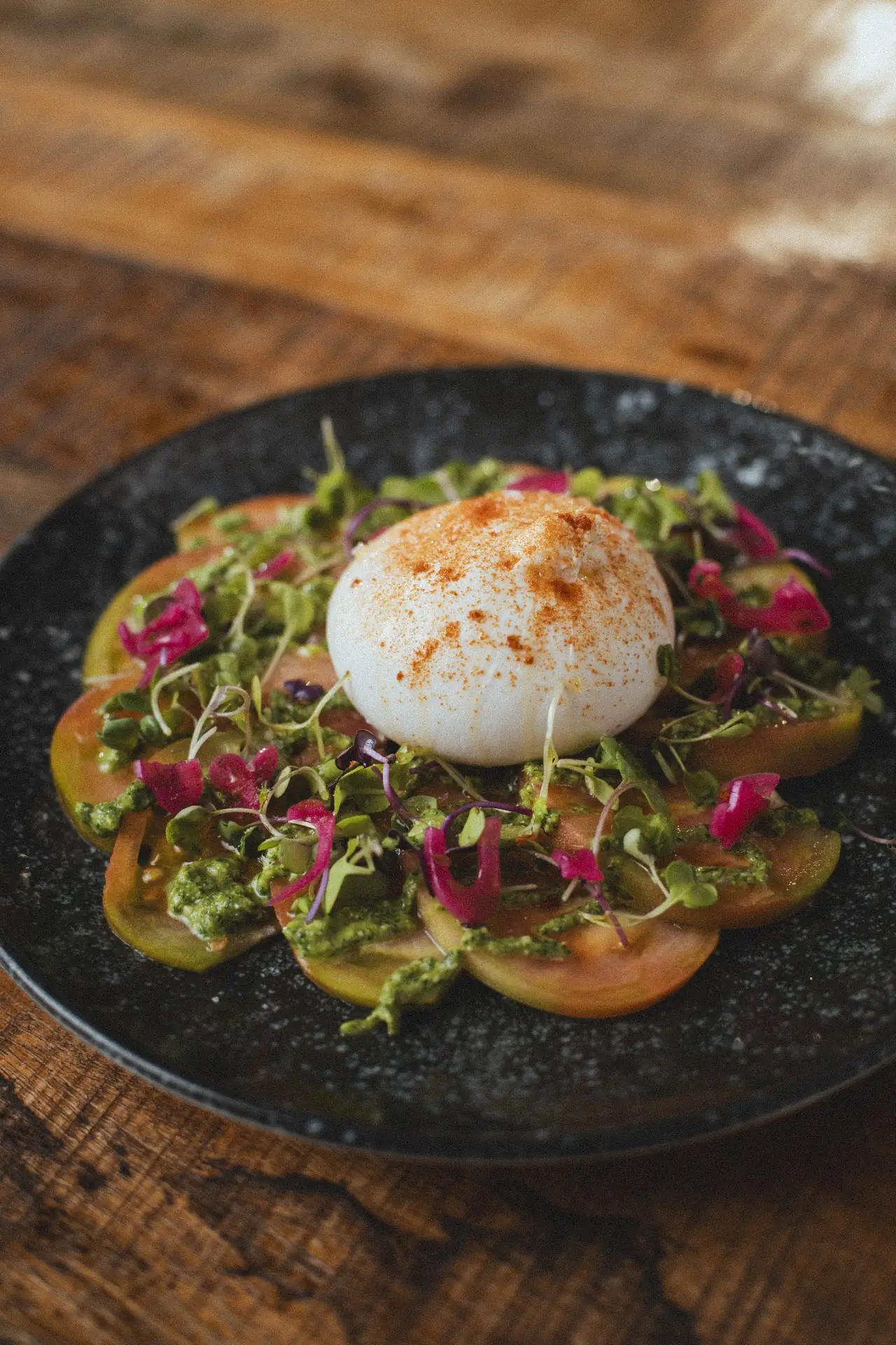 Round of white cheese over sliced heirloom tomatoes with greens and edible flower petals on a dark plate