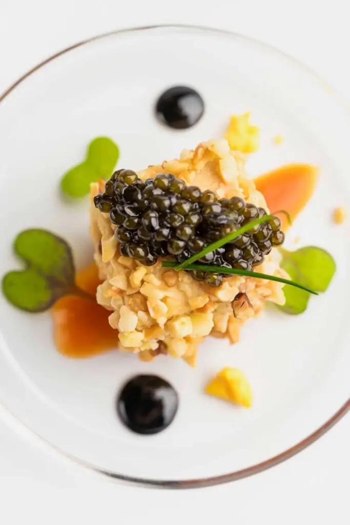 Overhead plated dish of a nutty tartare topped with black caviar and a chive, dotted with dark and amber sauces on a white plate