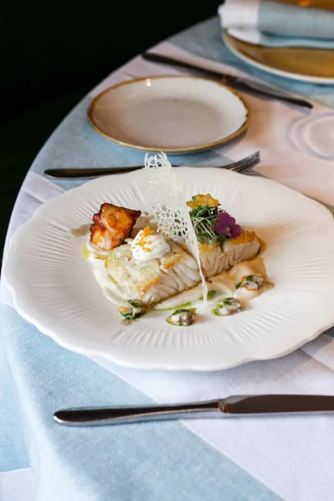 Plated white fish fillet in a pale sauce with a crisp lace tuile, an edible pansy flower and a seared cube, on a fluted white plate at a set table