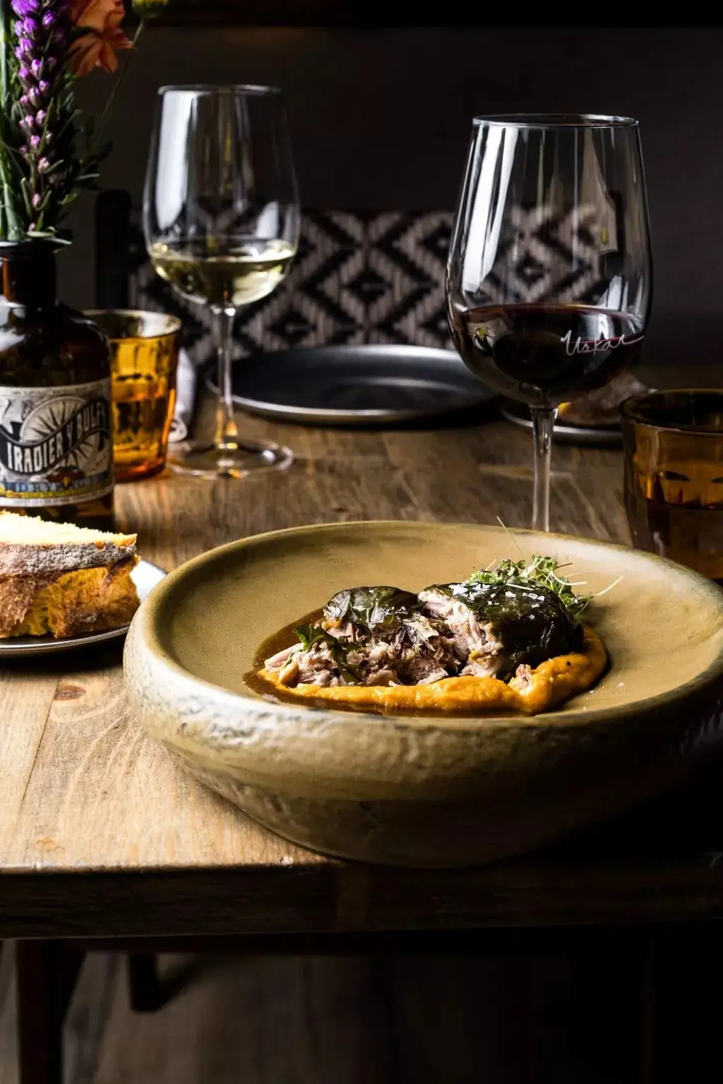 Rustic ceramic bowl of shredded braised meat wrapped in vine leaves over orange squash puree, with an 'Uskar'-etched glass of red wine, white wine and toasted bread on a wooden table