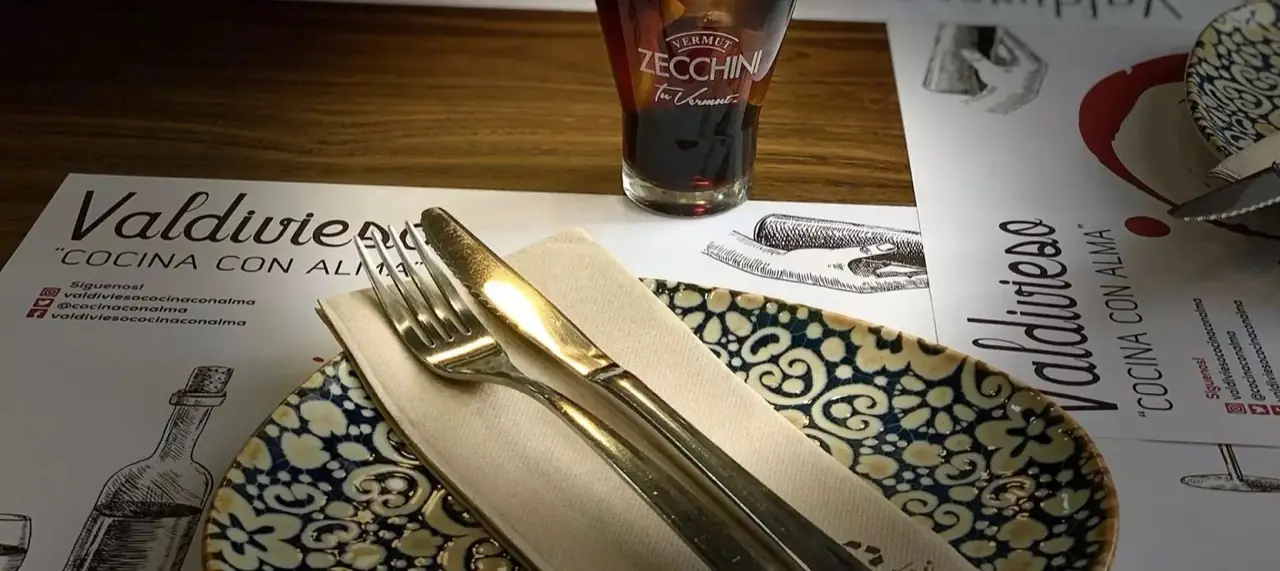 Table place setting on branded Valdivieso placemat with a patterned blue-and-white plate, folded napkin and cutlery, beside a tall glass of dark vermut served over ice