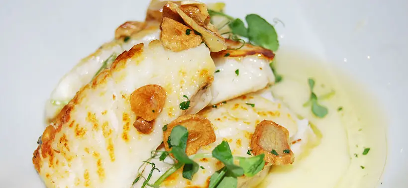 Pan-seared white fish fillets topped with crispy garlic slices and microgreens in a light broth, at Valentin Restaurant Xarcuteria