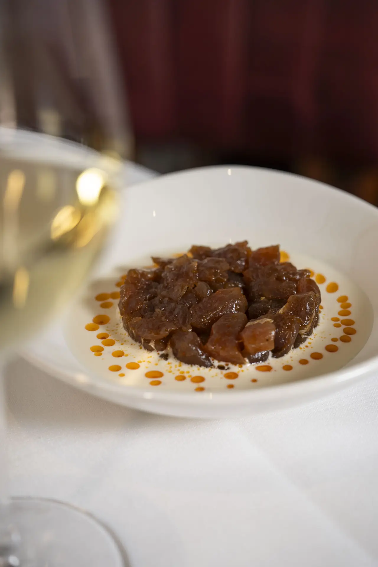 A shallow white bowl on a white tablecloth holding a round of glossy dark-red diced raw tuna, ringed by a neat circle of orange-tinted oil droplets, with a glass of white wine blurred in the foreground.