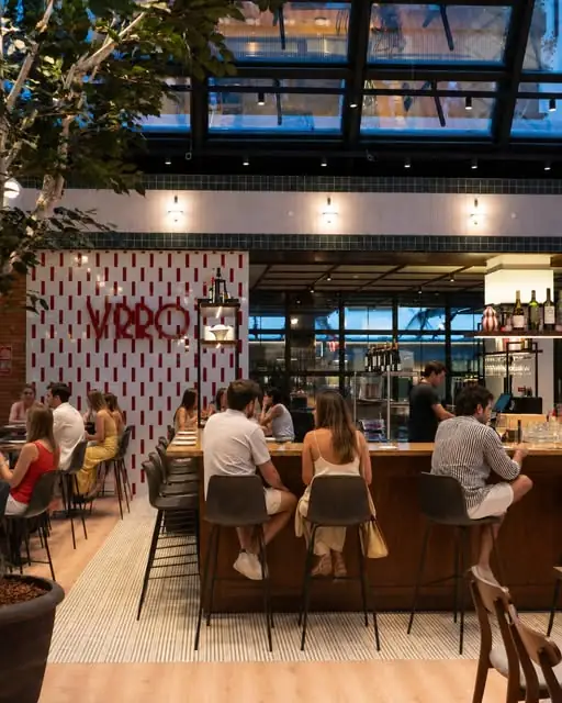 Interior dining room with patrons seated at a long wooden bar counter, red VRRO logo on a white tiled wall, hanging plants and a glazed roof