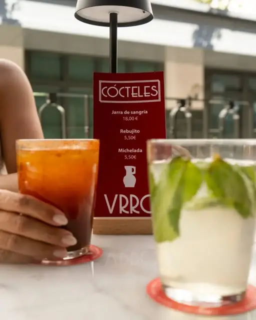 Two served drinks on a marble table, an orange sangria-style cocktail in a tumbler and a clear mint drink, with a red cocktails menu card behind