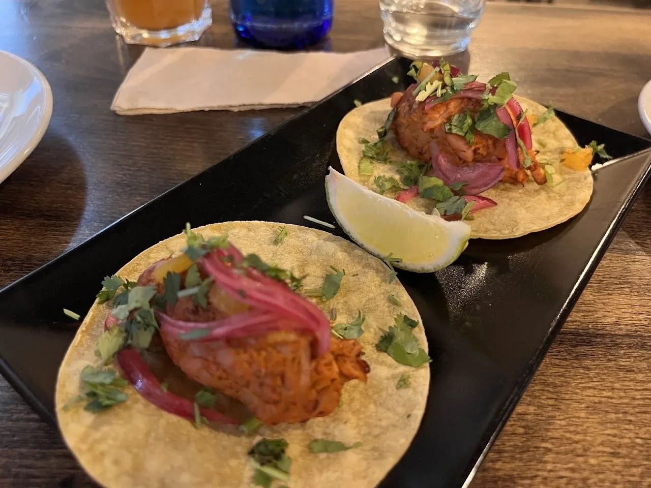 Two soft tacos on a black plate topped with pickled red onion, with a lime wedge