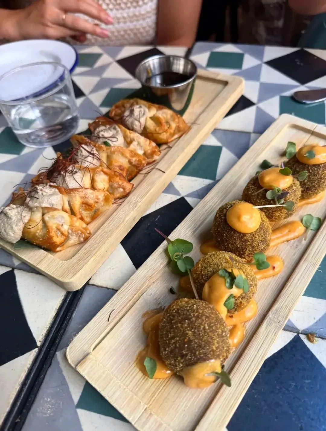 A wooden serving board of golden pastry rolls and round green-crusted croquettes on a tiled table