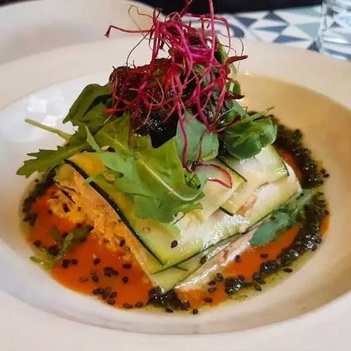 A plated stack of green courgette ribbons and salad leaves with a red-orange sauce