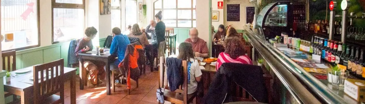 Interior dining room with diners at wooden tables and a bar counter lined with beer taps and bottles, faces blurred