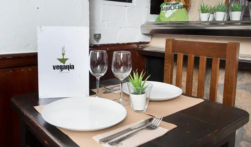 Set table with white plates, wine glasses and cutlery, and a Vegania Veggie Bar menu card standing against a white brick wall