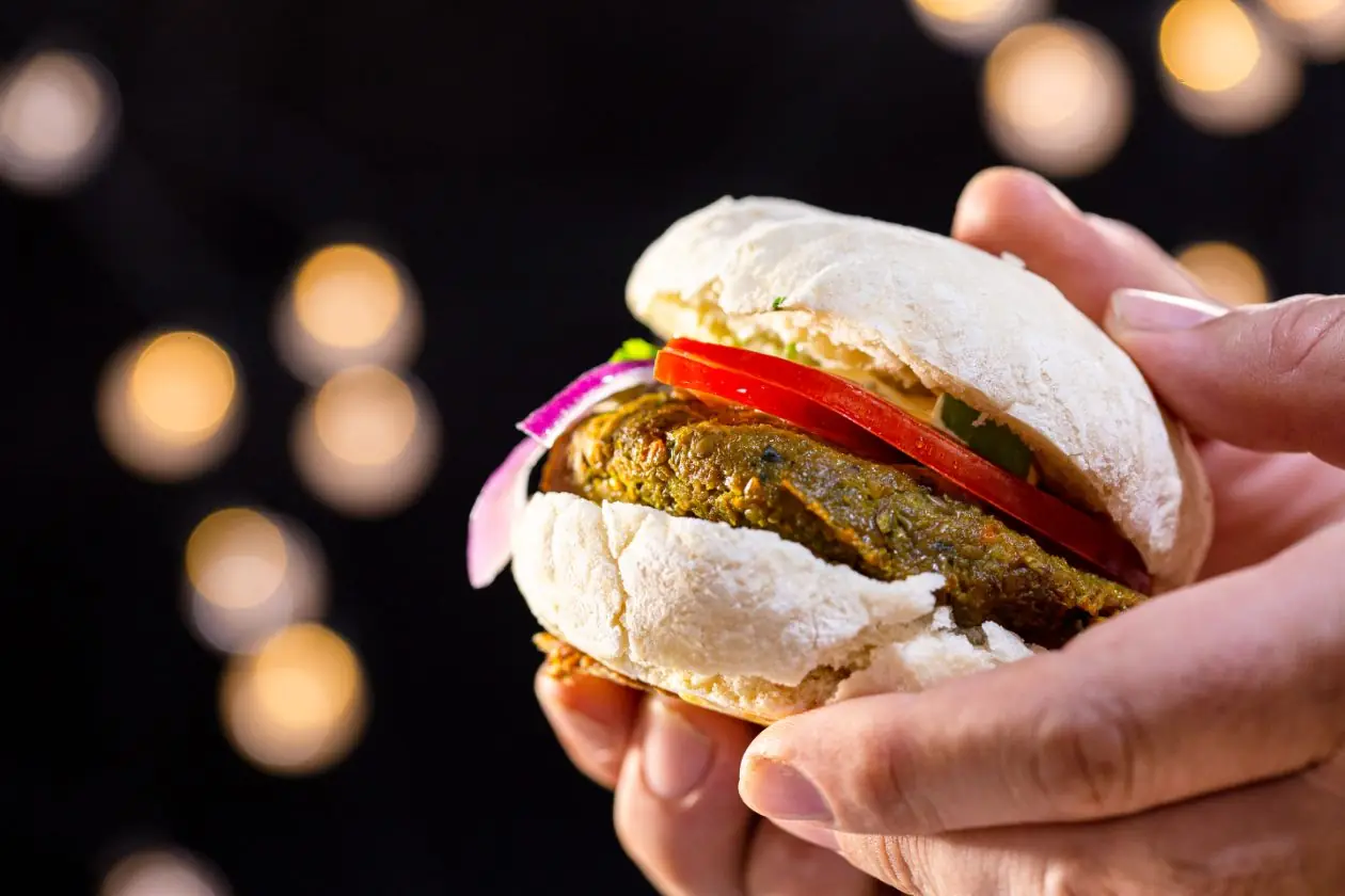 Hands holding a vegan burger with tomato and red onion in a soft white bun