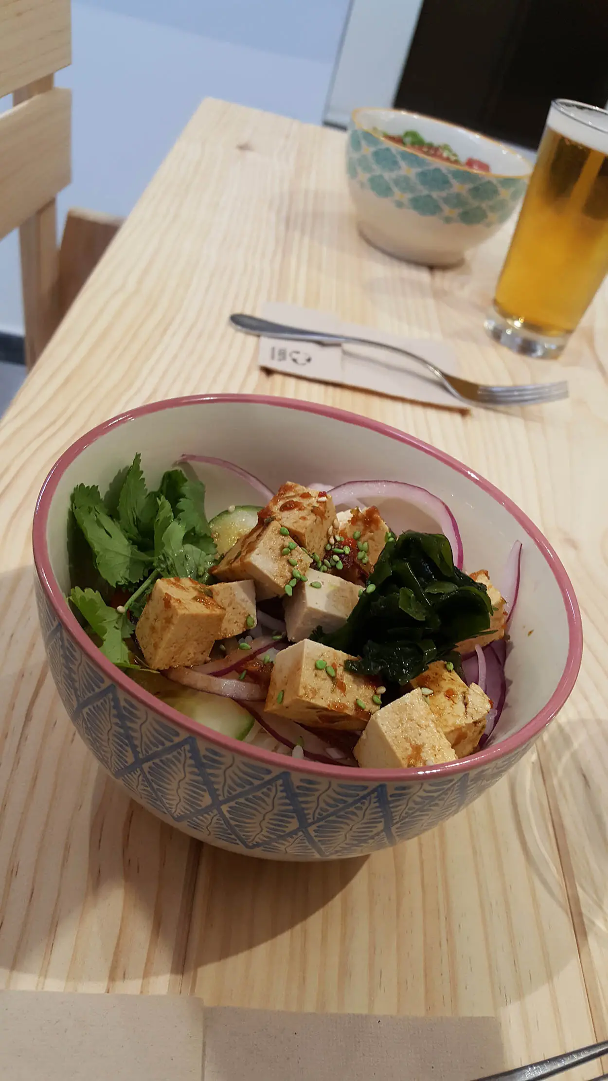 A patterned bowl of marinated tofu cubes with cucumber, red onion, cilantro and seaweed, with a beer in the background