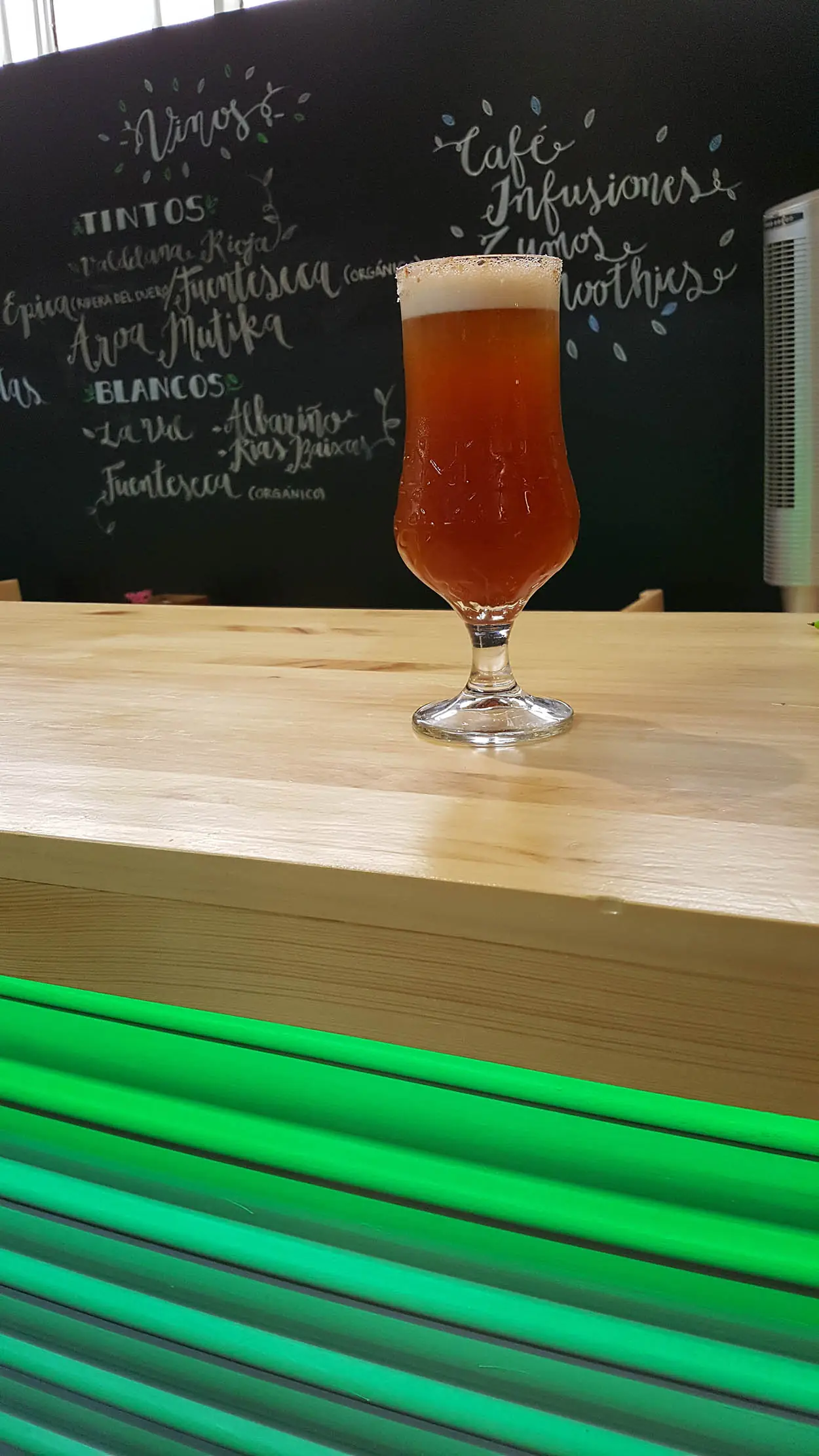 A footed glass of amber beer with a foam head on a wooden bar counter, chalkboard drinks menu behind
