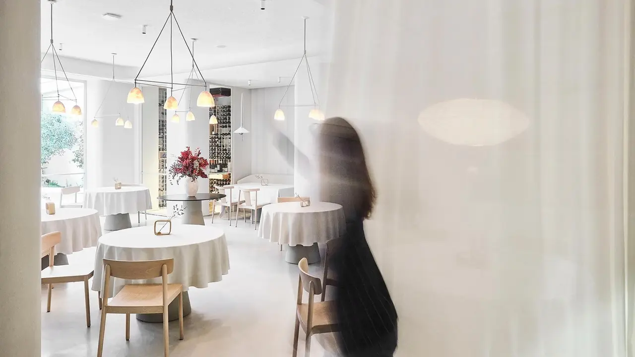 A bright white dining room of round tables in white cloths under alabaster pendant lamps, with a glass-fronted wine store at the back and a blurred figure passing