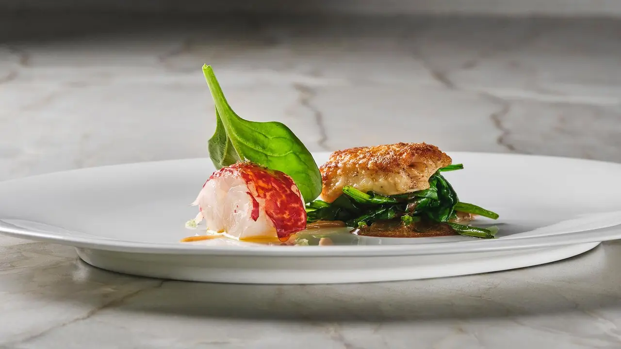 A piece of lobster tail and a golden-crusted fillet on wilted spinach with a spinach leaf, on a white plate over marble