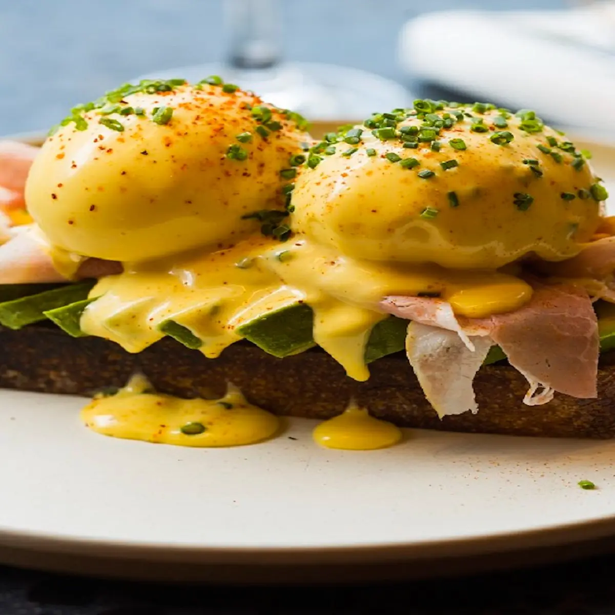 Two poached eggs Benedict with hollandaise sauce, chives, avocado, and ham on toasted bread at Veraz