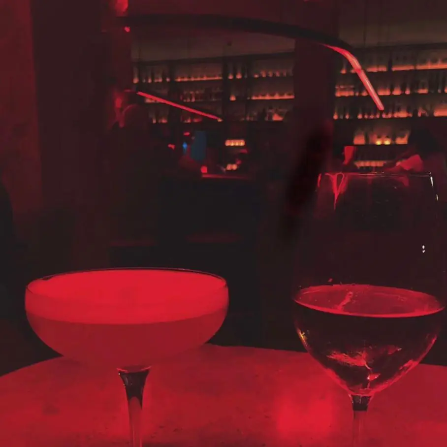 A coupe cocktail and a glass of red wine on a bar top under red lighting, with a backlit bottle-lined bar behind