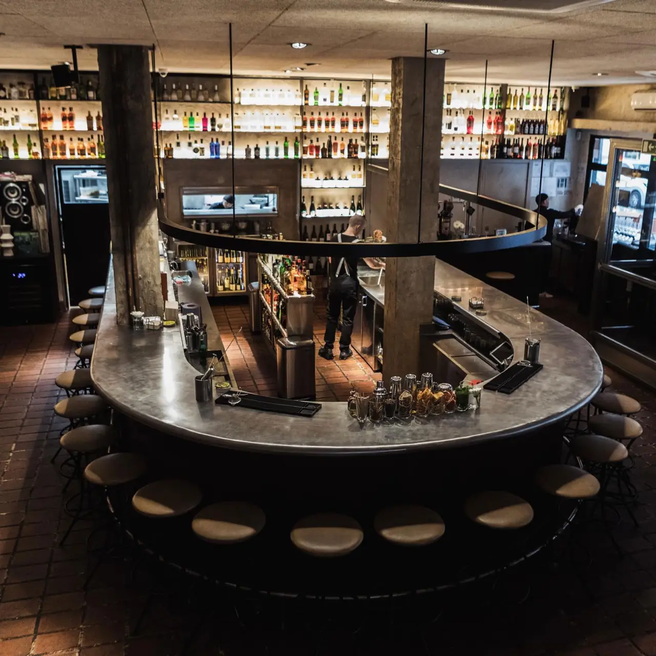 Large oval metal bar counter ringed with stools, a bartender working behind it and shelves of backlit bottles lining the walls