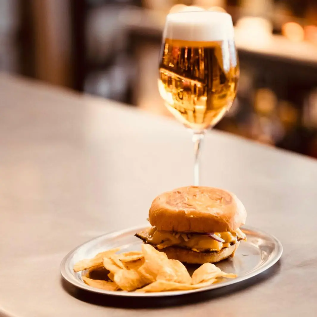 A grilled sandwich in a soft bun with potato crisps on a metal plate, beside a glass of golden beer on a bar top