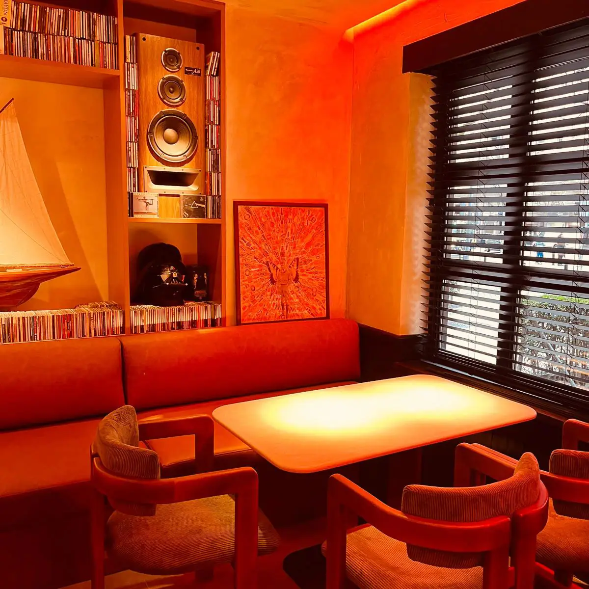 Warm-lit dining nook with an orange banquette, wooden armchairs and a table, shelves of CDs and a large hi-fi speaker, a model sailboat and framed art