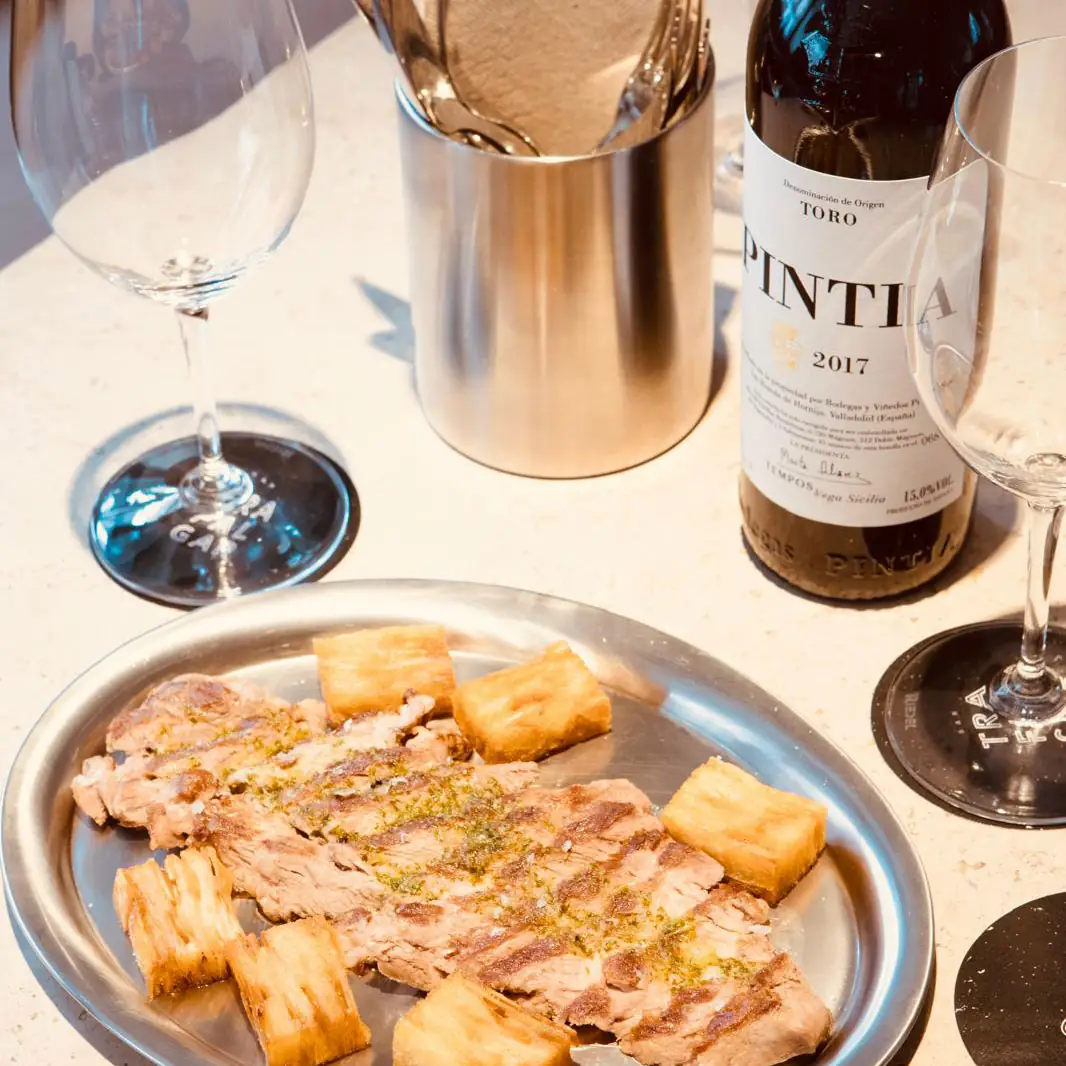 Sliced grilled meat with fried yuca on a metal plate, beside a bottle of Pintia Toro 2017 red wine and two wine glasses on a pale table
