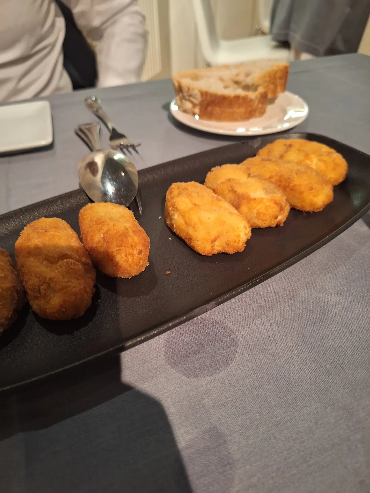 Row of breaded croquettes on a black tray with bread in background