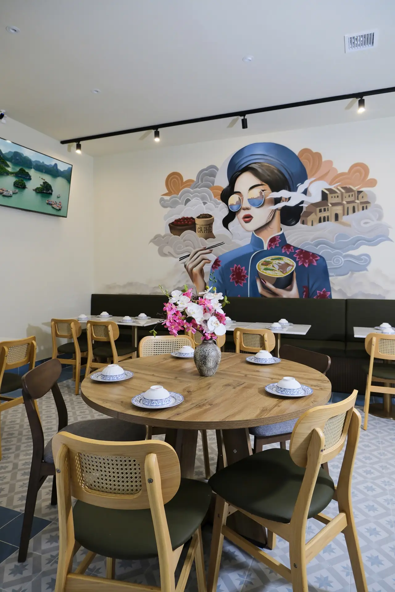 Vietnamese dining room with round wooden tables, cane-back chairs, patterned floor tiles and a large painted wall mural of a woman in an ao dai holding a bowl of pho