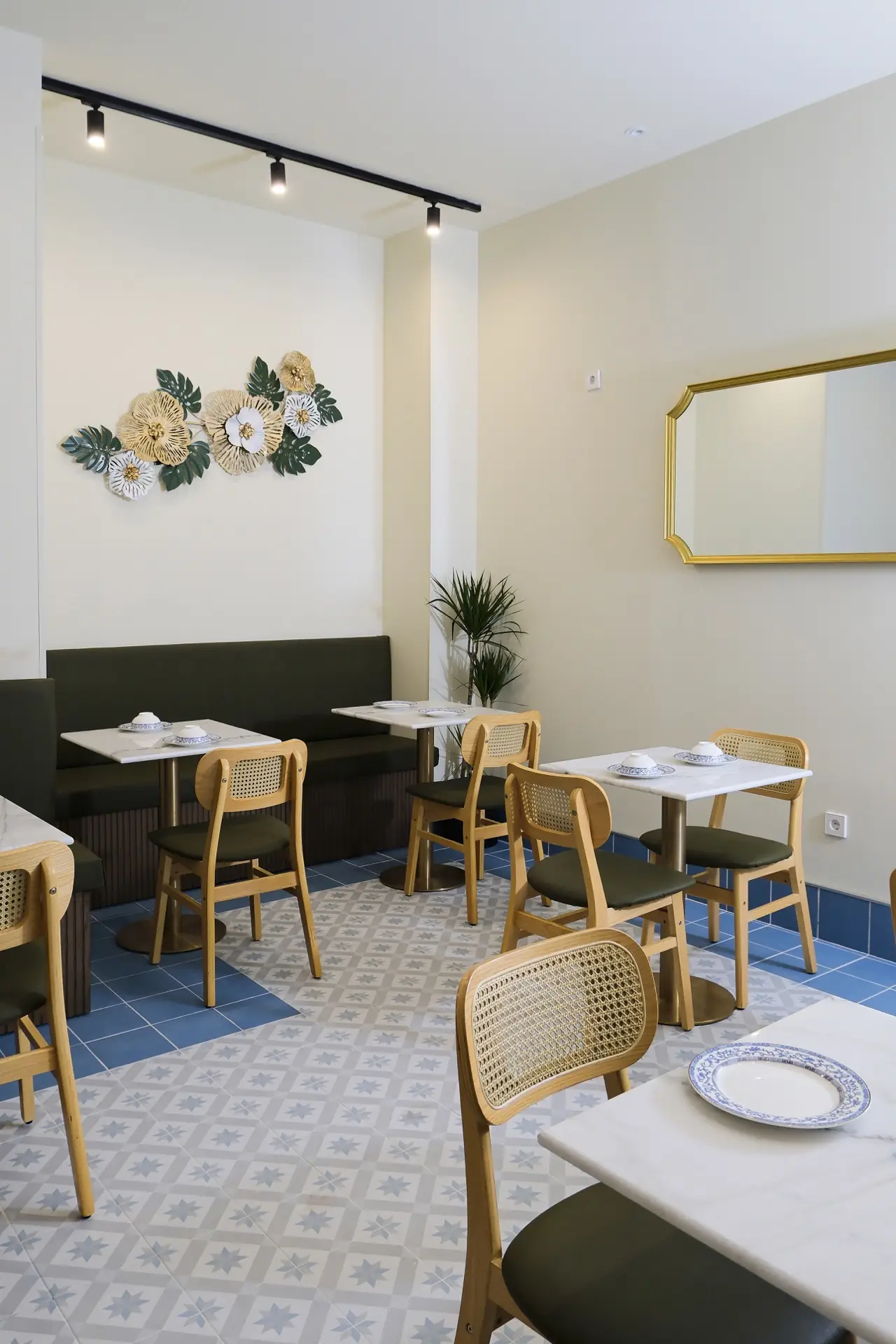 Bright dining room corner with marble-top tables, cane chairs, blue star-patterned floor tiles, a gold-framed mirror and a decorative metal-flower wall sculpture