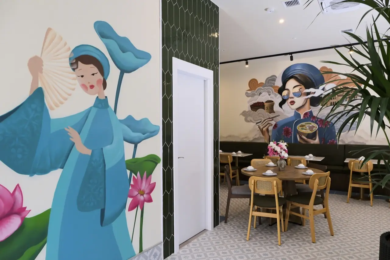 Interior view past a floor-to-ceiling mural of a woman with a fan into a dining room with round tables, cane chairs and a second pho-woman mural