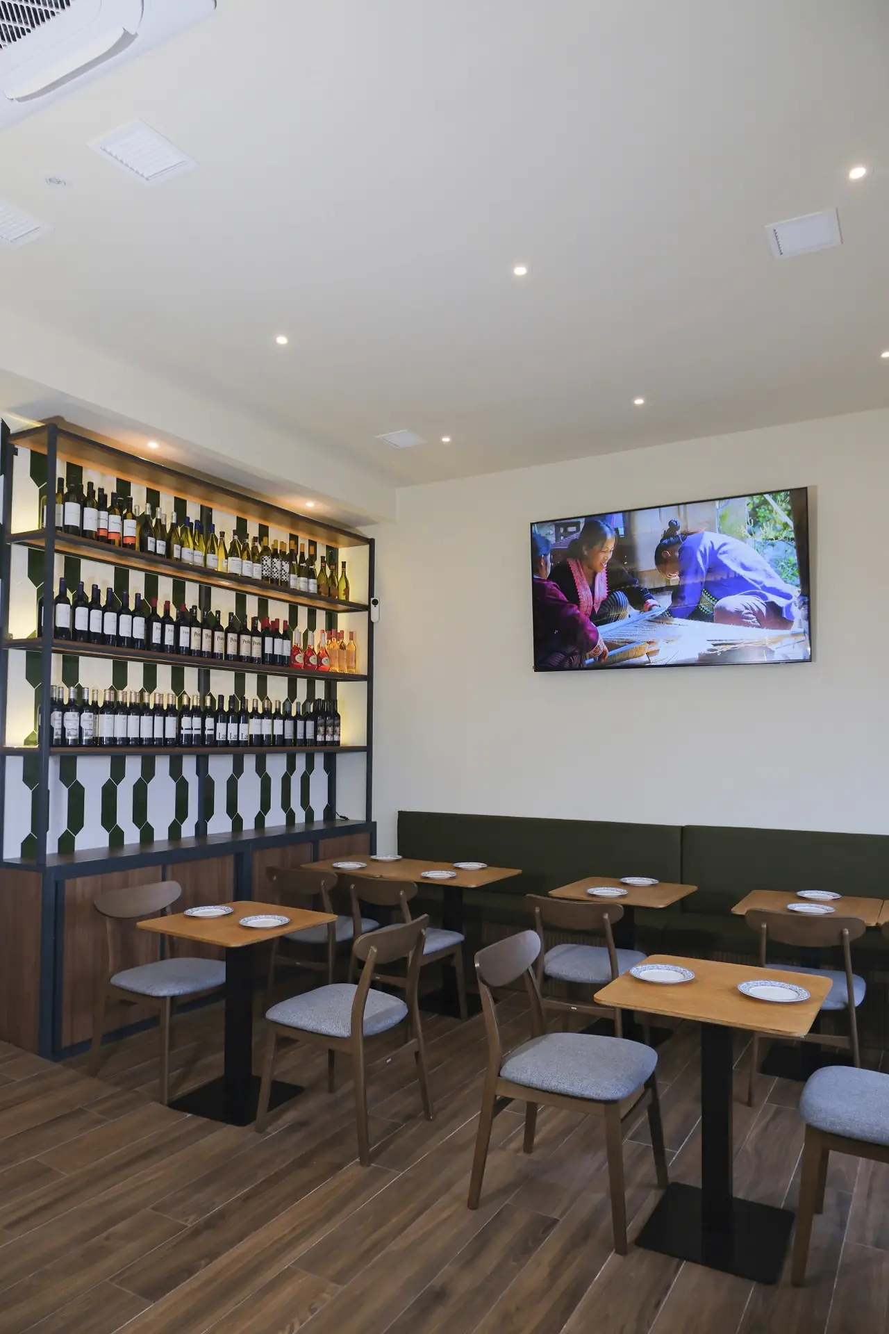 Dining area with a lit green-and-white tiled wine shelf full of bottles, wall-mounted TV, wooden tables and grey chairs on dark wood floor