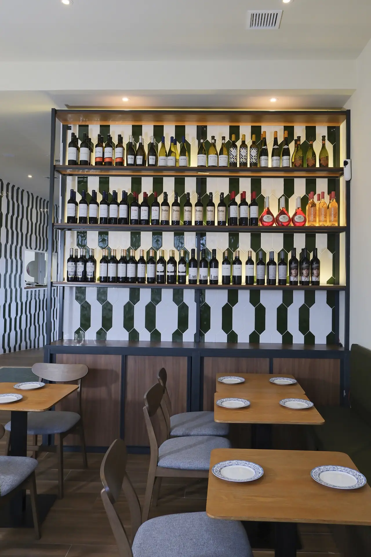 Dining room with a tall backlit wine display shelf on a green-and-white tiled wall and set wooden tables with blue-rimmed plates