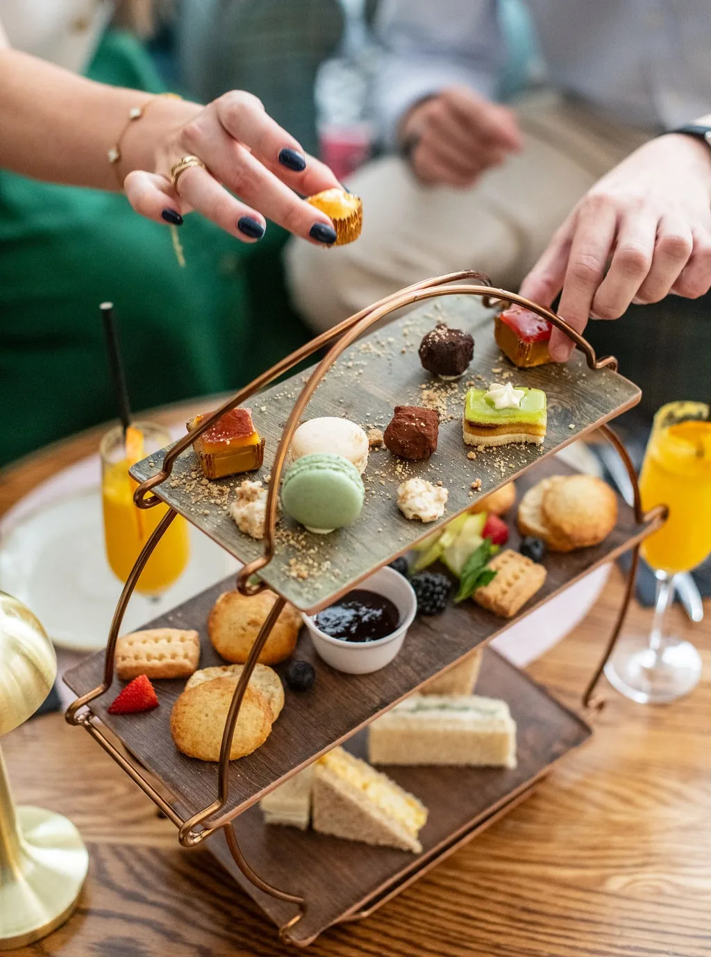 A tiered stand with an assortment of afternoon tea pastries, sandwiches, and sweets, with hands reaching for a piece
