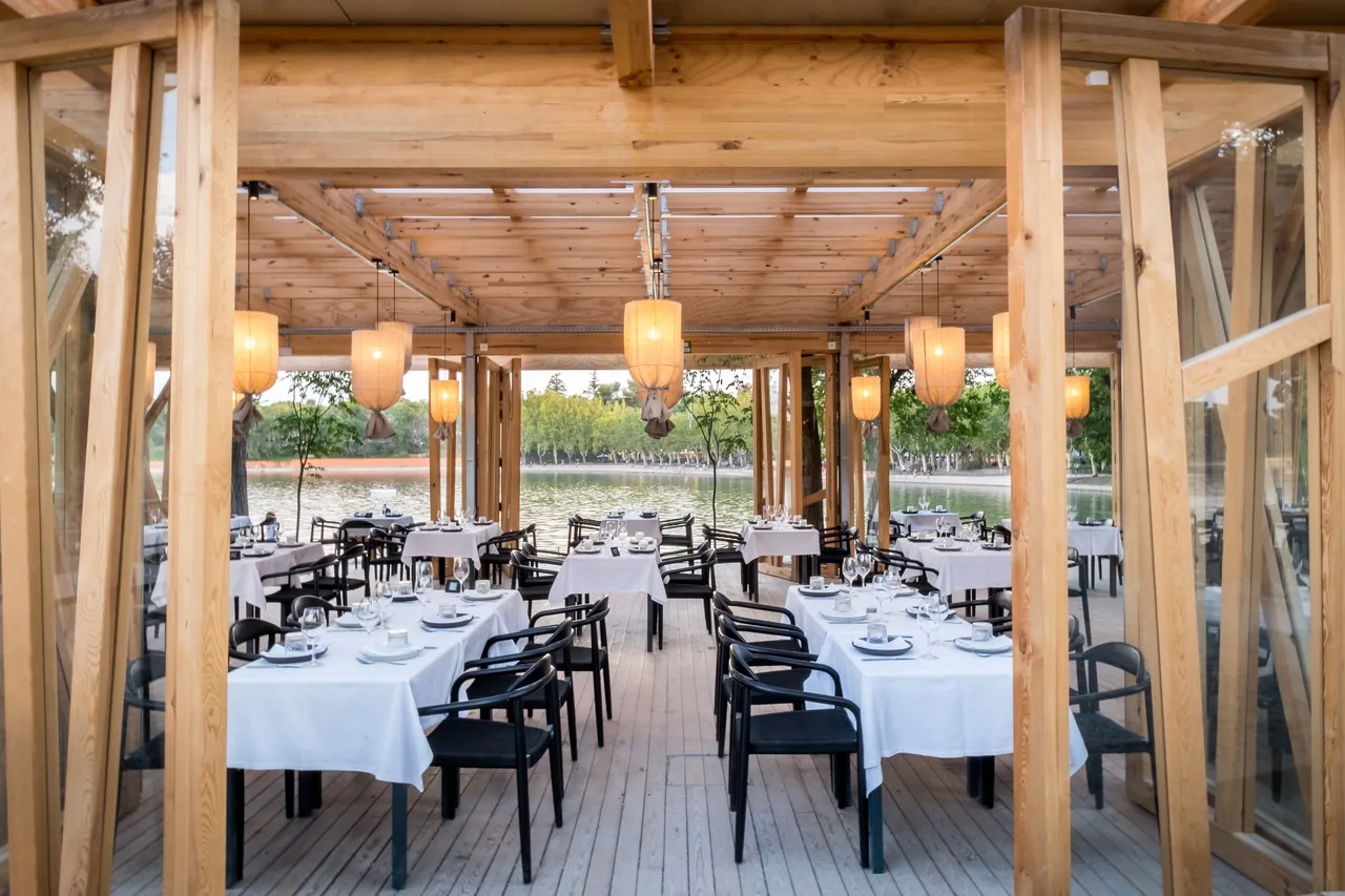 White-clothed tables under a timber pavilion with paper lanterns, looking out over a lake