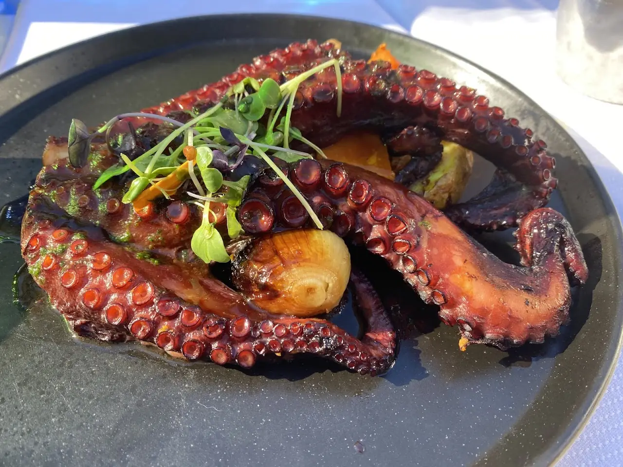 Grilled octopus tentacles with charred onion and microgreens on a black plate.