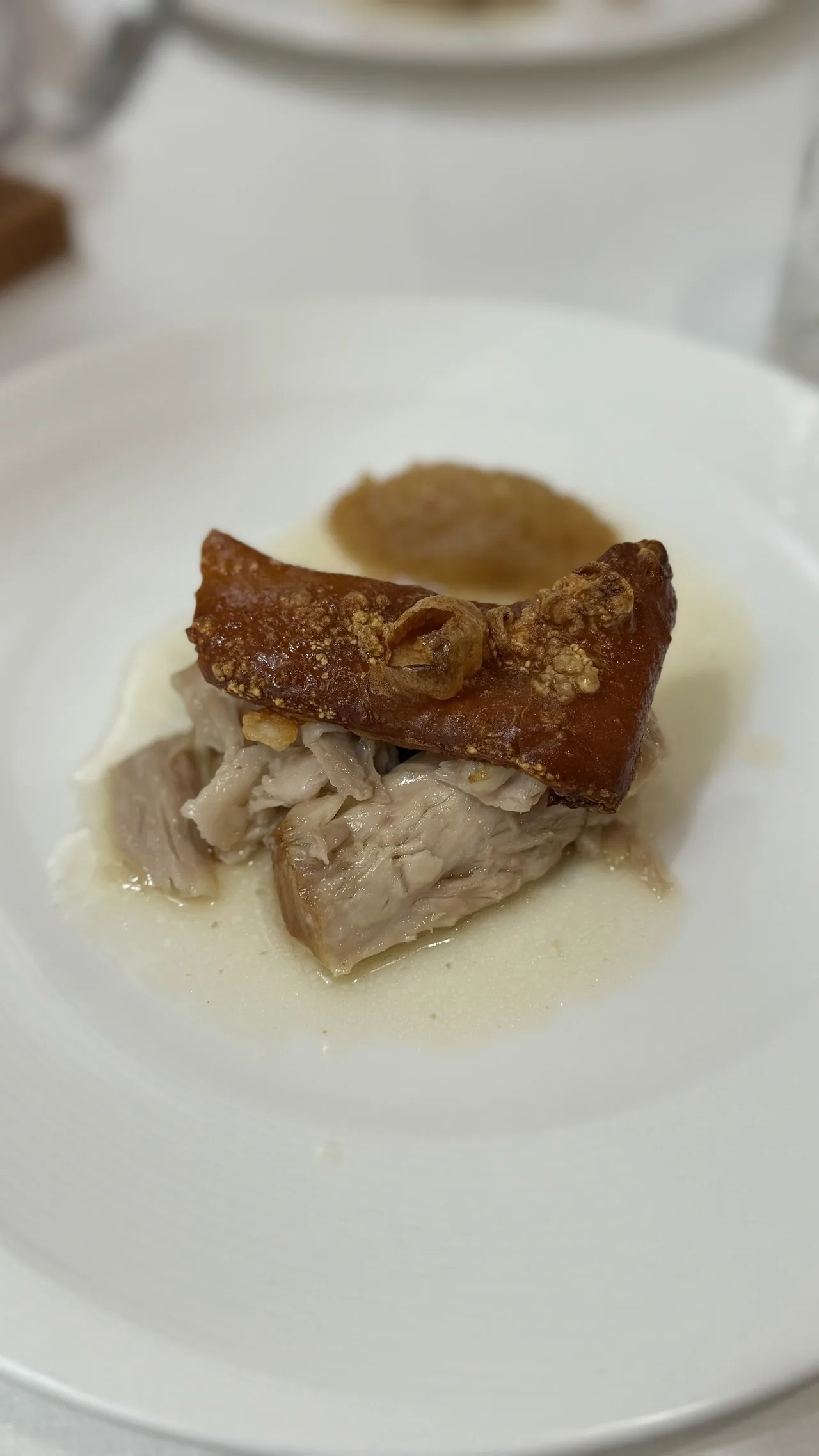 Shredded roast pork topped with a piece of golden crackling on a white plate, with a spoonful of purée behind