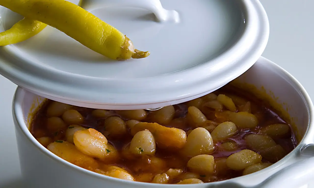 White bean stew in a covered ceramic pot with a pickled yellow pepper on the lid